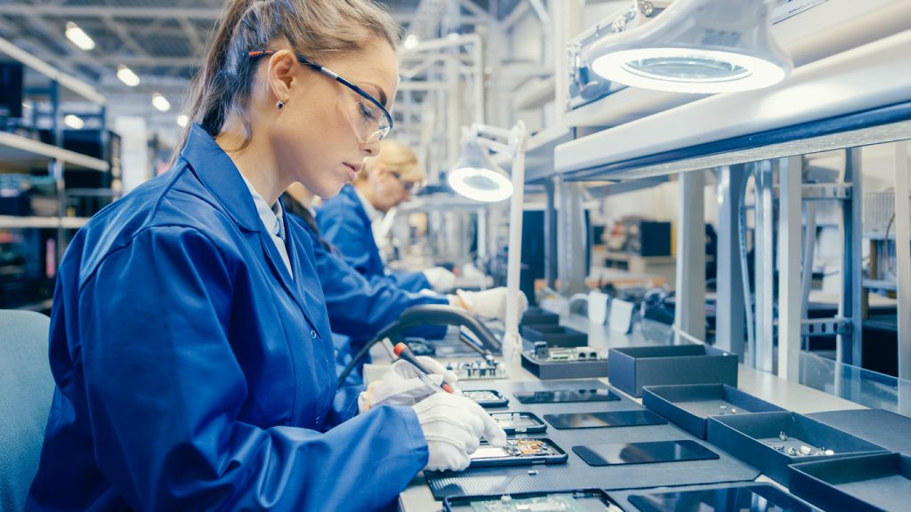 Woman Electronics Factory Worker in Blue Work Coat and Protective Glasses is Assembling Smartphones with Screwdriver. High Tech Factory Facility with more Employees in the Background.