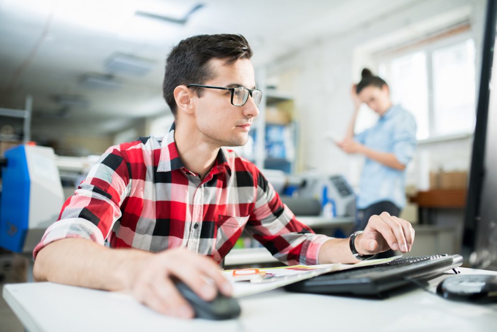 Serious young printing specialist in glasses sitting at computer and working on design project in office