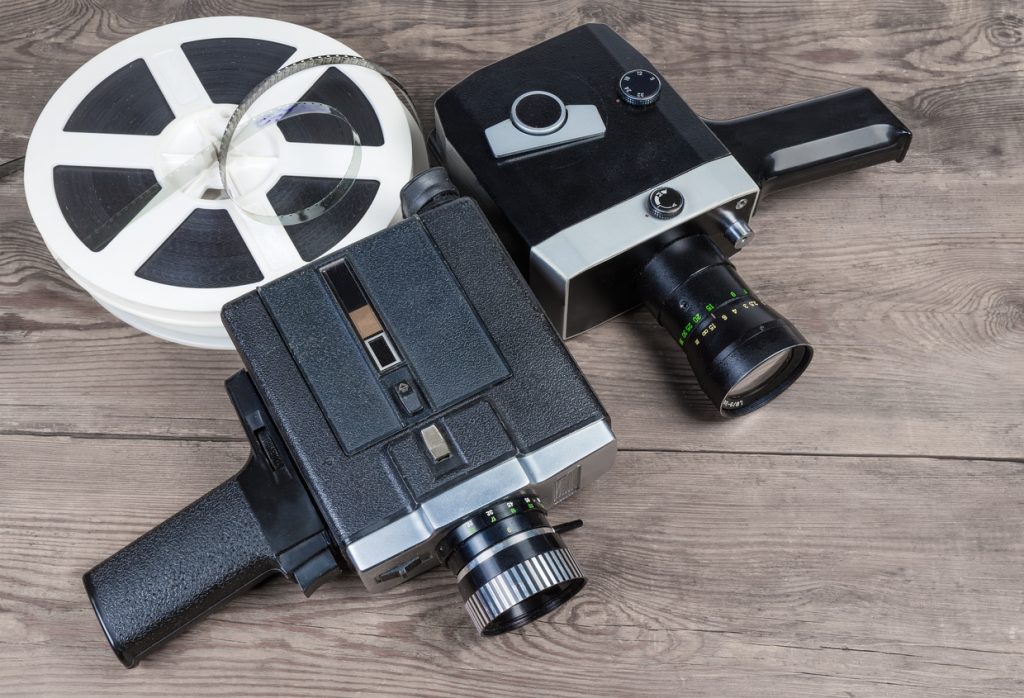 Two old vintage amateur film movie cameras powered by clockwork motor and electric motor and reels of motion picture films Super 8mm format on an old wooden surface