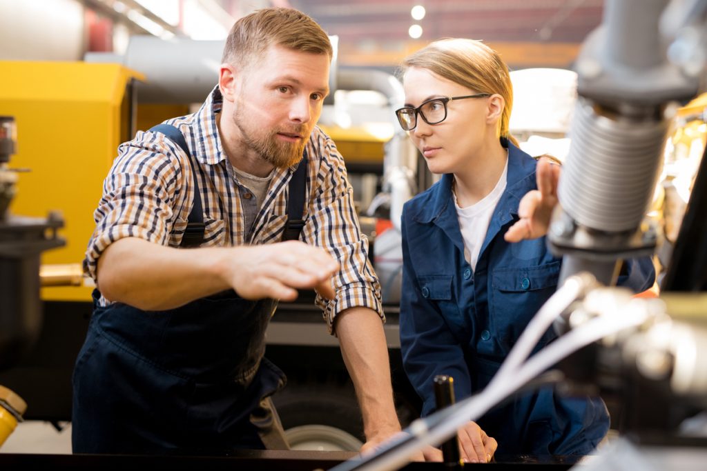 One of young confident technicians pointing at mechanism of industrial equipment during discussion of its qualities with colleague (One of young confident technicians pointing at mechanism of industrial equipment during discussion of its qualities wit